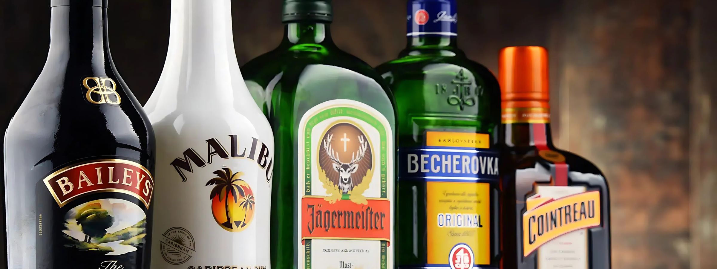 Assorted liqueur bottles (cream, fruit, herbal) with glasses, dessert dishes and cocktail tools on a bar surface