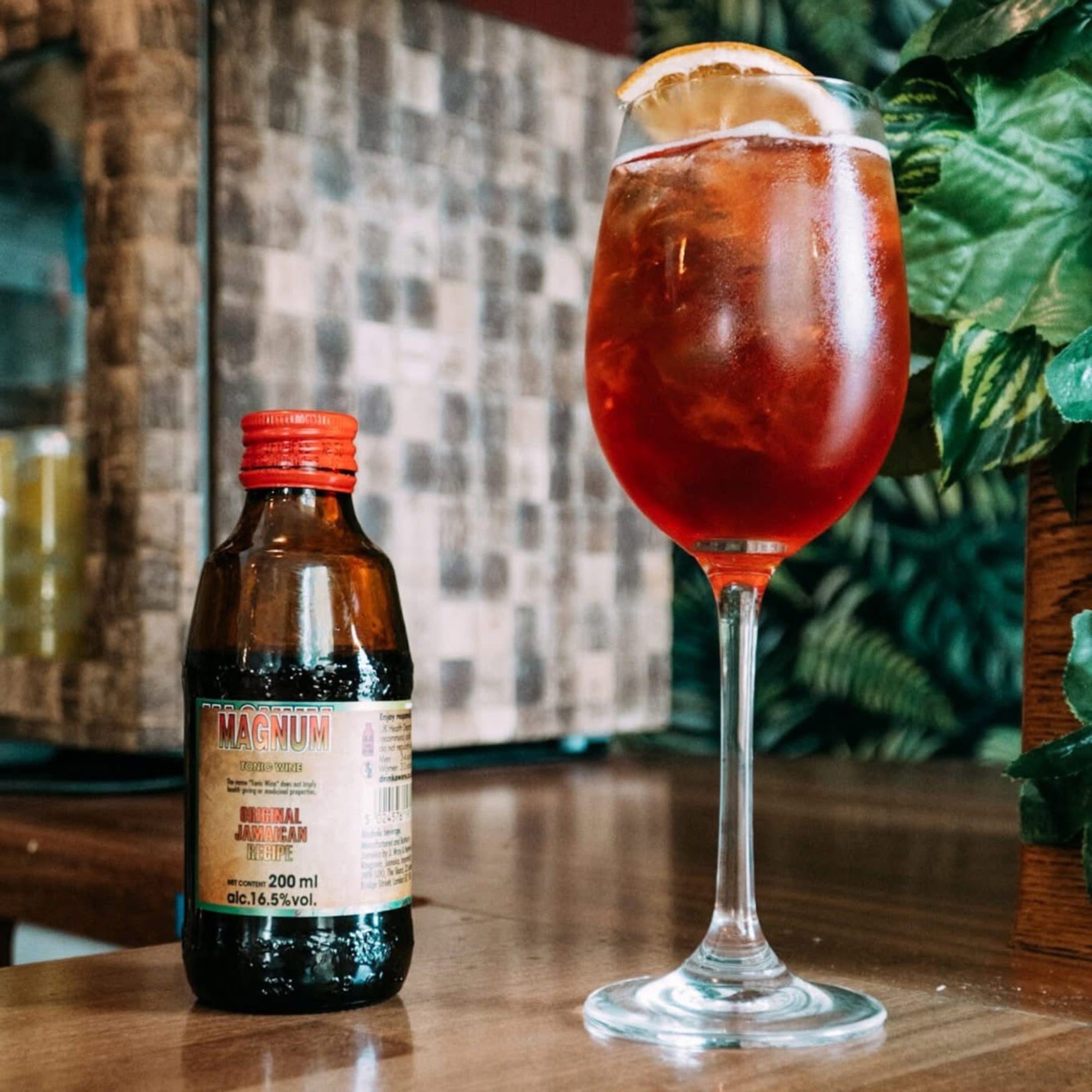 Bottle of Magnum Jamaican rum and a glass of red cocktail with a lemon wedge on a wooden surface.